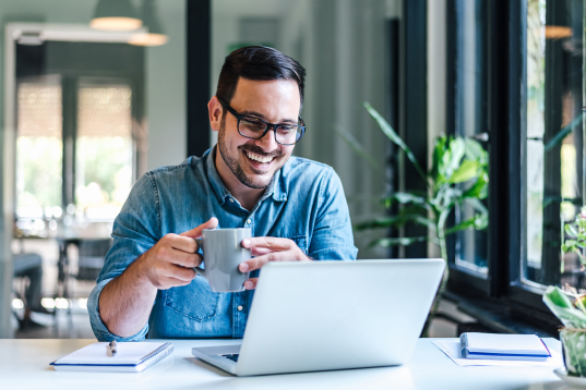 Business man drinks coffee while smiling at his laptop 