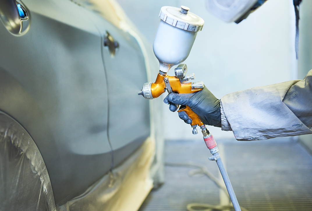 A person in protective clothing applying paint to a car. Only the person’s hand and arm are shown.