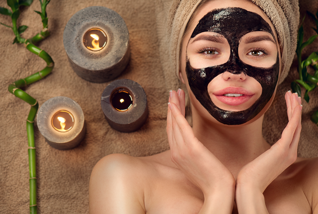 A woman with a dark face mask lies on a bath towel. A bath towel is wrapped around her head. Next to her are three candles. A woman with a dark face mask lies on a bath towel. A bath towel is wrapped around her head. Next to her are three candles.