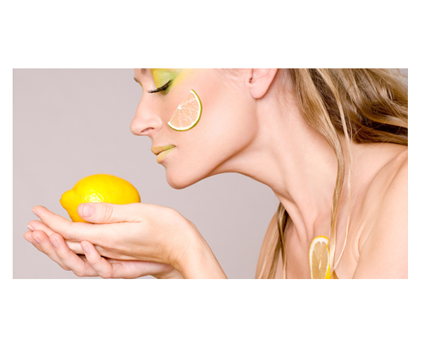 Eine Frau mit gelb geschminkten Lippen und Lidern und einer Zitronenscheibe auf der Wange riecht an einer Zitrone, die sie in den Händen hält.