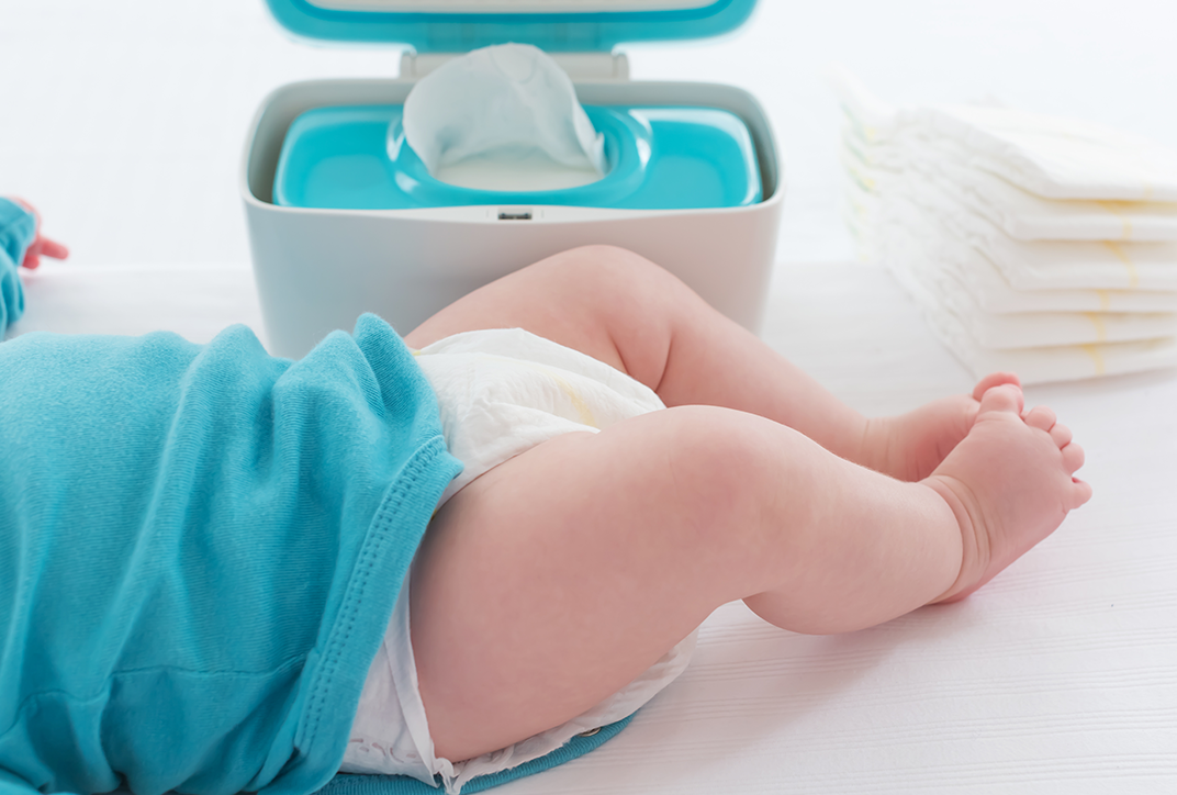 A baby on a changing table. In the background is a box of wet wipes, with a pile of diapers next to it.