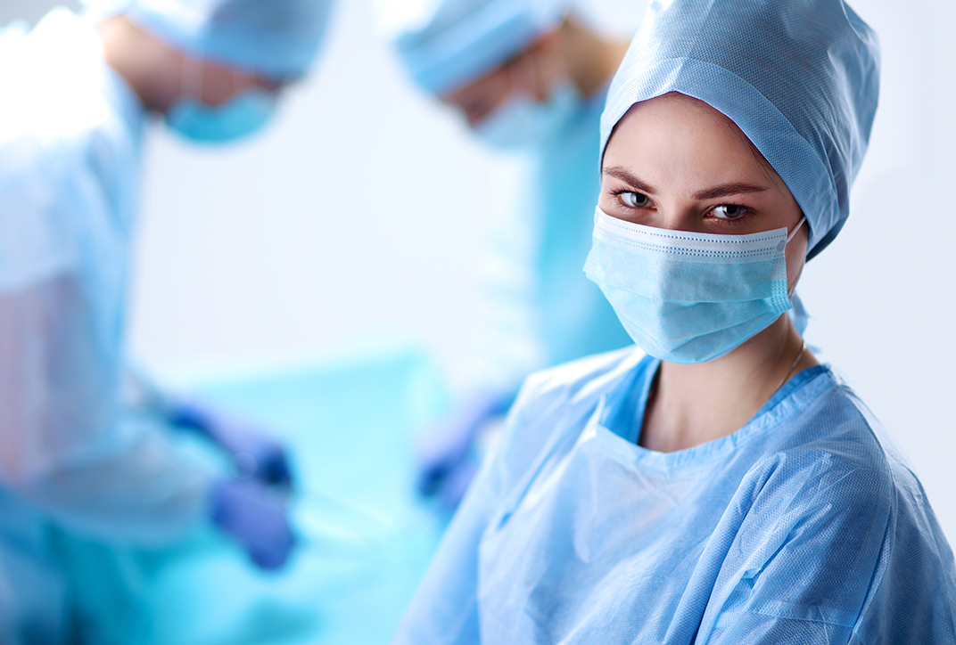 A lady in an operating gown and mask. In the background are two more people in operating gowns.